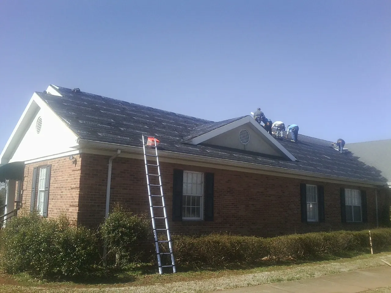 Crew tearing shingles off a brick ranch roof with a ladder set — Metal Roof Repair work by our crew in Foster
