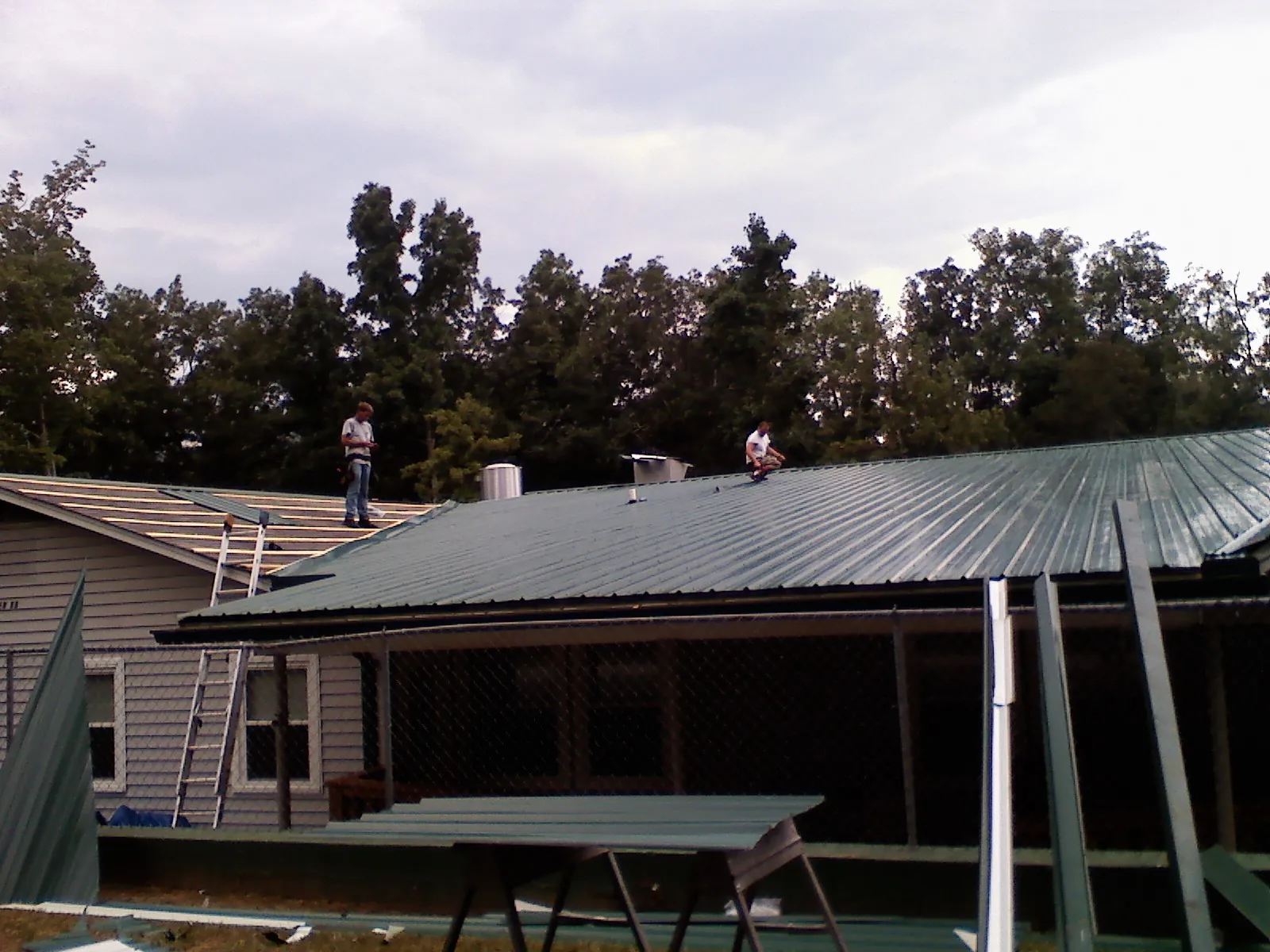 Two roofers installing green metal panels above a covered porch in Foster